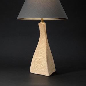 Könnte beinhalten: Eine beige Tischlampe aus Keramik mit einer strukturierten Oberfläche und einem schwarzen Stoffschirm.