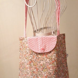 Puede incluir: Una bolsa de mano de tela de color rosa y blanco con estampado floral, con un bolsillo de color rosa y blanco con estampado geométrico y un botón rosa. La bolsa tiene una correa de color rosa y blanco con estampado.