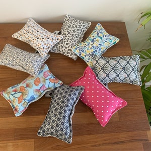 Può includere: Una collezione di piccoli cuscini rettangolari in tessuto con vari motivi, tra cui triangoli, floreali e disegni geometrici. I cuscini sono in tonalità di blu, rosa, bianco e arancione. Sono disposti su una superficie di legno.