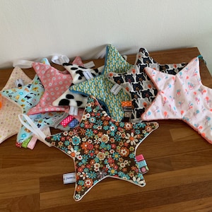 May include: A collection of eight star-shaped baby toys made of soft fabric. The stars are in various colors and patterns, including pink, blue, yellow, brown, and white. Each star has a small tag attached to it with the word "Pummel" printed on it.
