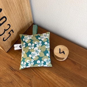 Puede incluir: Un peque&ntilde;o saquito de tela rectangular con fondo verde azulado y un estampado de flores blancas y hojas de color verde claro. Incluye una trabilla de tela a cuadros verdes y blancos en la parte superior y una peque&ntilde;a etiqueta lateral blanca.