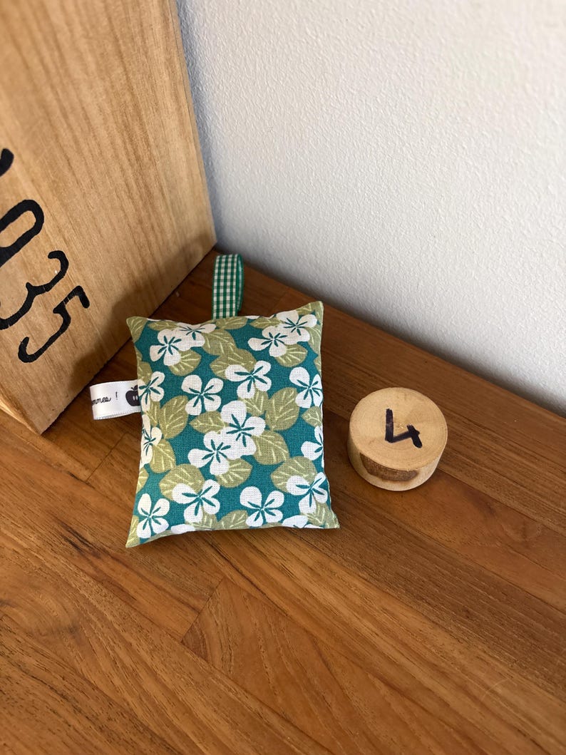 Puede incluir: Un peque&ntilde;o saquito de tela rectangular con fondo verde azulado y un estampado de flores blancas y hojas de color verde claro. Incluye una trabilla de tela a cuadros verdes y blancos en la parte superior y una peque&ntilde;a etiqueta lateral blanca.