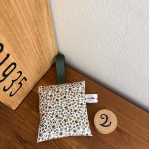 Puede incluir: Un saquito de tela cuadrado con un estampado floral peque&ntilde;o en tonos verde, marr&oacute;n y blanco. Incluye una cinta verde oscuro en la parte superior y una peque&ntilde;a etiqueta blanca lateral con el texto 4 p'tites p. Un disco de madera redondo muestra el n&uacute;mero 2.