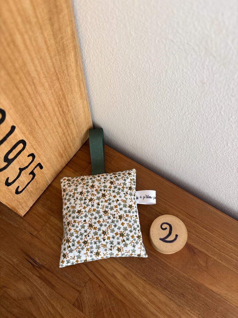 Puede incluir: Un saquito de tela cuadrado con un estampado floral peque&ntilde;o en tonos verde, marr&oacute;n y blanco. Incluye una cinta verde oscuro en la parte superior y una peque&ntilde;a etiqueta blanca lateral con el texto 4 p'tites p. Un disco de madera redondo muestra el n&uacute;mero 2.