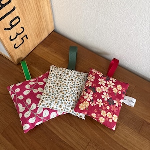 Puede incluir: Tres peque&ntilde;os saquitos rectangulares de tela con cintas para colgar. El saquito de la izquierda es rosa con patrones de hojas blancas y una cinta verde. El saquito central presenta un peque&ntilde;o estampado floral con una cinta verde oscuro. El saquito de la derecha es rojo con patrones florales y una cinta roja.