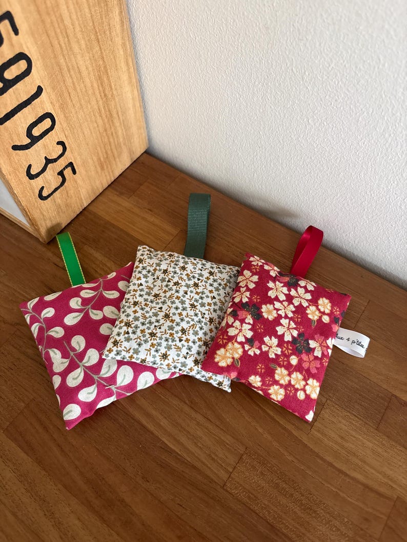 Puede incluir: Tres peque&ntilde;os saquitos rectangulares de tela con cintas para colgar. El saquito de la izquierda es rosa con patrones de hojas blancas y una cinta verde. El saquito central presenta un peque&ntilde;o estampado floral con una cinta verde oscuro. El saquito de la derecha es rojo con patrones florales y una cinta roja.