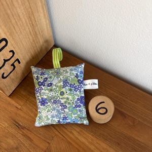 Puede incluir: Un peque&ntilde;o saquito de tela cuadrado con un estampado floral en tonos azules, verdes y blancos. Tiene un lazo de cinta a rayas verdes y blancas en la parte superior y una etiqueta de tela blanca en el lateral con el texto Aux 4 p'tites.