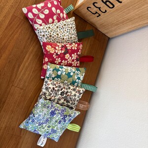 Puede incluir: Seis saquitos de tela cuadrados y peque&ntilde;os con diversos estampados florales y de hojas en tonos rosa, crema, rojo, azul verdoso, gris y azul. Cada saquito incluye una peque&ntilde;a trabilla de tela en un lado, uno con una etiqueta blanca con texto.