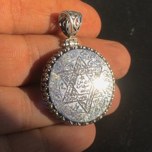 Könnte beinhalten: Silberanhänger mit kreisförmigem Design. Der Anhänger zeigt einen sechszackigen Stern in der Mitte, umgeben von komplizierter Schrift und einem dekorativen Rand. Der Bügel ist verziert, mit einem filigranen Design.