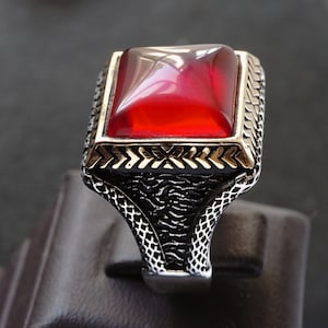 May include: A silver ring featuring a large, square, red gemstone set in an intricate, ornate design.