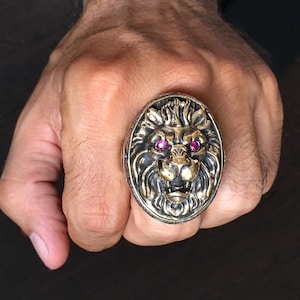 May include: A large, oval-shaped ring featuring a detailed lion's head with ruby-colored eyes. The ring has a gold-colored lion's head set against a darker, possibly oxidized, metal band. The lion's mane and facial features are intricately carved.