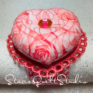 May include: A red heart-shaped fabric napkin ring with a pink floral pattern and a sparkly pink gem in the center. The ring is trimmed with red lace.