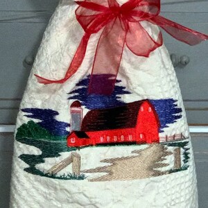 Puede incluir: Calcetín de Navidad de tela blanca con un lazo de cinta rojo y una imagen bordada de un granero rojo con un cielo azul y hierba verde.