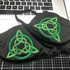 May include: Two black face masks with green embroidered Celtic knot designs. The knots are in the shape of a circle with a triangle inside.