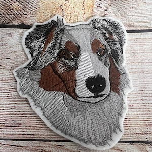 May include: Embroidered patch of an Australian Shepherd dog's head. The dog has a white and brown coat with blue eyes. The patch is made of white felt and has a stitched border.