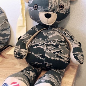 May include: A camouflage-patterned teddy bear with a brown nose and black eyes. The bear is wearing a small American flag patch on its foot.