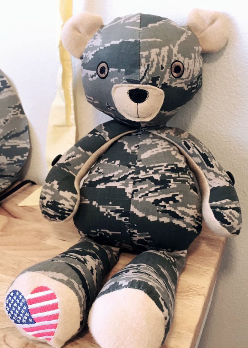 May include: A camouflage-patterned teddy bear with a brown nose and black eyes. The bear is wearing a small American flag patch on its foot.