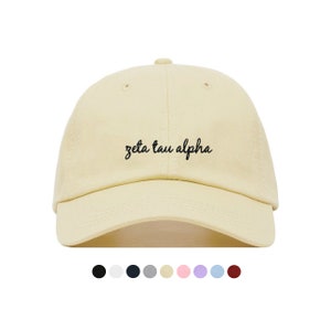 Puede incluir: Una gorra de béisbol de color amarillo claro con texto bordado en negro que dice "zeta tau alpha".