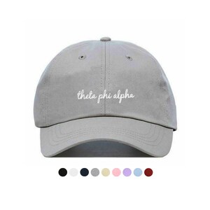 Puede incluir: Gorra de béisbol gris claro con el texto "theta phi alpha" en escritura blanca en la parte delantera. La gorra tiene una visera curva y una corona redondeada. Se muestran opciones de color debajo de la gorra.