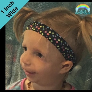 May include: A young girl with blonde hair is wearing a black headband with rainbow and white stars. The headband is 1 inch wide. The text "THE BAHA BOWTIQUE" is visible in the image.