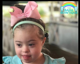 Pode incluir: Uma menina pequena com um laço rosa e uma faixa de cabeça verde claro. A faixa de cabeça é um design simples sem enfeites. A menina está vestindo uma camisa azul claro. A imagem tem um logotipo no canto superior direito que diz "THE BAHA BOWTIQUE".