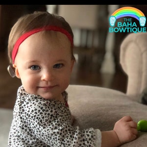 May include: A young child wearing a red headband and a white and black animal print shirt. The child is sitting on a light brown chair. The child is smiling and looking at the camera. The text "THE BAHA BOWTIQUE" is visible in the upper right corner of the image.