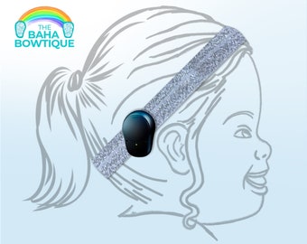 May include: A silver glitter headband with a black device attached, positioned on a child's head. The illustration features a smiling child's profile. The logo "The Baha Bowtique" is in the upper left corner, with a rainbow and feet.