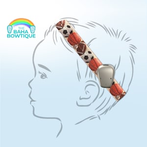May include: A headband with a sports ball pattern, including footballs and basketballs, paired with a silver hearing device. The headband is designed to secure the device. The logo "The Baha Bowtique" is in the upper left corner.