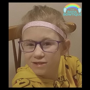 May include: A person wearing a light pink glitter headband and purple-framed glasses. The person is wearing a yellow hooded sweatshirt. The image includes the text "THE BAHA BOWTIQUE" with a rainbow graphic.
