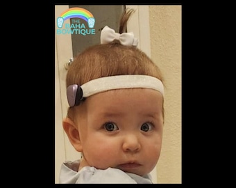 Pode incluir: Um bebê usando uma faixa branca com um pequeno laço branco e um acessório roxo. O bebê tem cabelo castanho e está olhando para a câmera. O logotipo "THE BAHA BOWTIQUE" está no canto superior esquerdo.