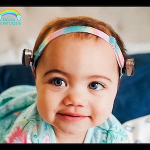 May include: A young child wearing a headband with a rainbow tie-dye pattern. The headband is holding a hearing aid on each side of the child's head. The child is wearing a light blue and pink floral print shirt.