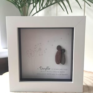May include: A white framed picture with a grey background and a quote about a daughter. The quote reads "A Daughter is someone you laugh with, dream with and love with all your heart". The picture also features a stone art piece of two people holding each other.