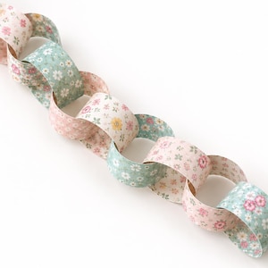 May include: A decorative paper chain composed of interlocking circular links. The links feature alternating floral patterns in soft shades of pink, white, and sage green, with small blossoms and foliage printed on a light-colored background.
