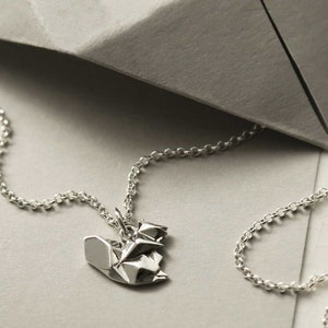 May include: A silver necklace with a folded paper origami pendant. The pendant is shaped like a heart and has a textured, crumpled look.