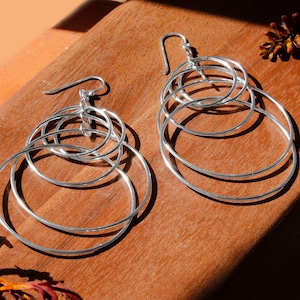 Może przedstawiać: Para srebrnych kolczyków koła z wieloma koncentrycznymi okręgami. Kolczyki leżą na drewnianej powierzchni.