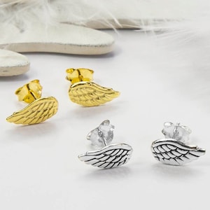 May include: Four pairs of stud earrings in the shape of wings. Two pairs are gold-colored, and two are silver-colored. The earrings are displayed on a white surface with a feather-like background.