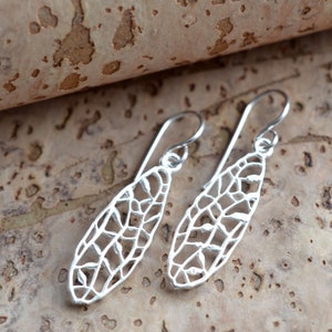 May include: A pair of silver-colored, openwork earrings. Each earring has a long, slender, teardrop shape with a delicate, web-like design. The earrings hang from simple, curved ear wires, and are displayed on a textured, brown surface.