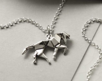 May include: A silver origami horse pendant necklace. The horse pendant, with its geometric design, hangs from a fine chain. The necklace is presented on a grey surface, highlighting the jewellery's intricate details and design.