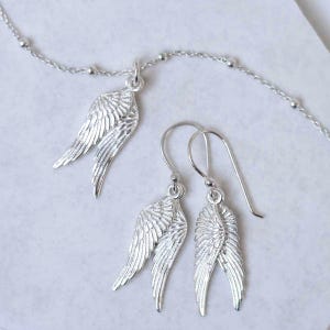 May include: A silver jewellery set featuring a necklace pendant and earrings, all in a wing design. The necklace has a delicate chain with small beads. The earrings have a hook closure. The wings have detailed feather textures.