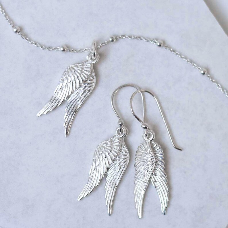 May include: A silver jewellery set featuring a necklace pendant and earrings, all in a wing design. The necklace has a delicate chain with small beads. The earrings have a hook closure. The wings have detailed feather textures.
