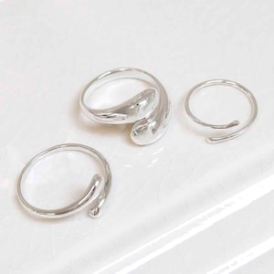 May include: Three silver rings with distinctive designs. One ring features a large, curved form, while the other two have a minimalist, open design. The rings are presented on a white surface, highlighting their polished, reflective surfaces.