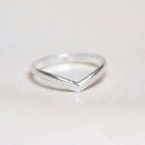 Op de afbeelding: Een minimalistische zilveren ring met een V-vormig ontwerp. De ring is gemaakt van gepolijst metaal en ligt op een witte ondergrond. De eenvoudige, geometrische vorm van de ring maakt het een veelzijdig accessoire.