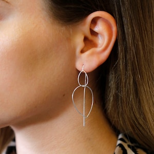 May include: Silver dangle earrings with a geometric design featuring two connected circles and a long, thin rod.