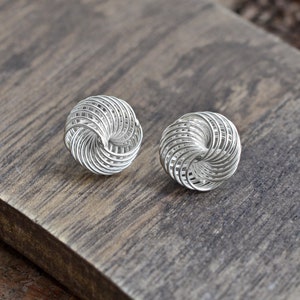 May include: A pair of silver stud earrings. Each earring is a sphere constructed from tightly wound, thin metal wires, forming a woven, open design. The earrings are displayed on a wooden surface.