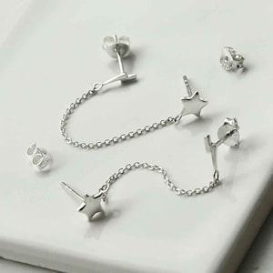 Puede incluir: Un par de pendientes de estrella plateados con una cadena que los conecta. Los pendientes están diseñados para ser usados en ambos lóbulos de las orejas.