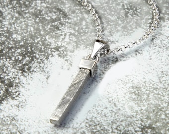 May include: A silver necklace with a long, thin pendant that resembles a sliver of metal. The pendant has a textured surface and is attached to a silver chain.