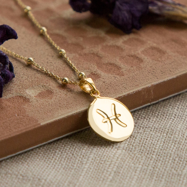 May include: Gold-colored necklace with a pendant featuring the Pisces zodiac symbol. The pendant is oval-shaped and hangs from a delicate chain with small, round beads. The necklace is displayed on a brown surface.