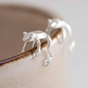 May include: Two silver frog-shaped cup clips, one in focus, clinging to the rim of a cream-coloured mug. The frogs have detailed features and are positioned as if climbing. The mug's rim is a dark brown.