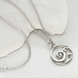 Puede incluir: Un collar plateado con un colgante circular con un dise&ntilde;o de ola arremolinada. El colgante est&aacute; unido a una delicada cadena. El collar se muestra sobre una superficie blanca.