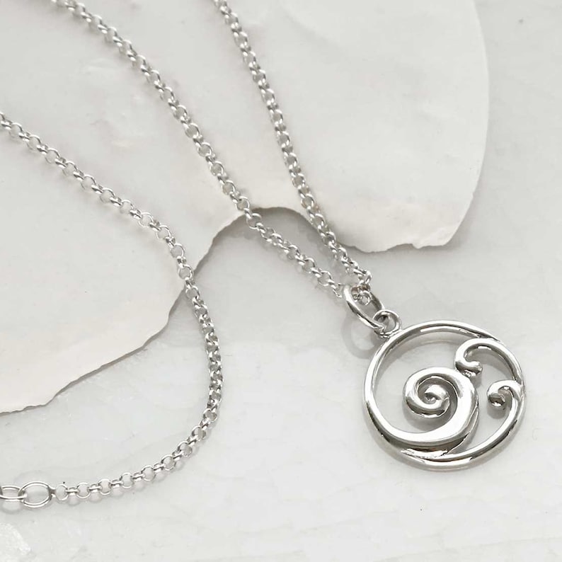 Puede incluir: Un collar plateado con un colgante circular con un dise&ntilde;o de ola arremolinada. El colgante est&aacute; unido a una delicada cadena. El collar se muestra sobre una superficie blanca.